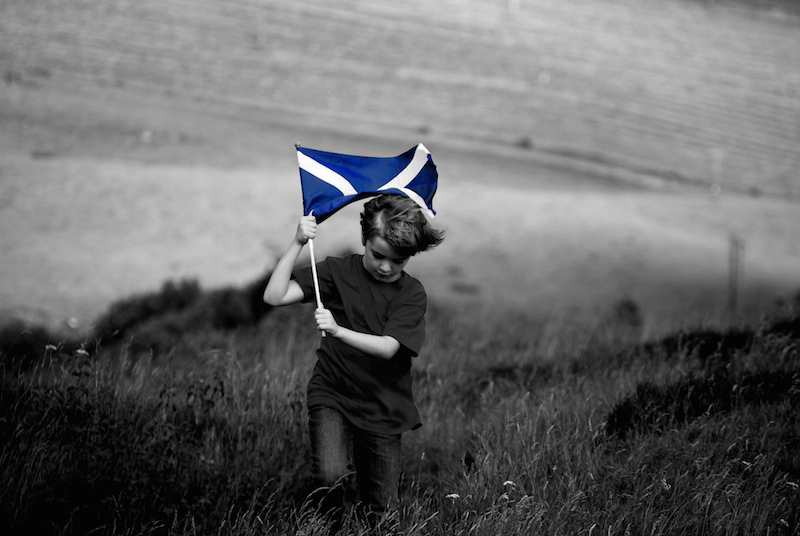 a black and white photo of a boy running through a field carrying a Scottish flag, which is coloured