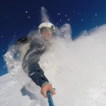 a deep blue sky behind a man snowboarding and kicking up a lot of fresh snow powder into his face