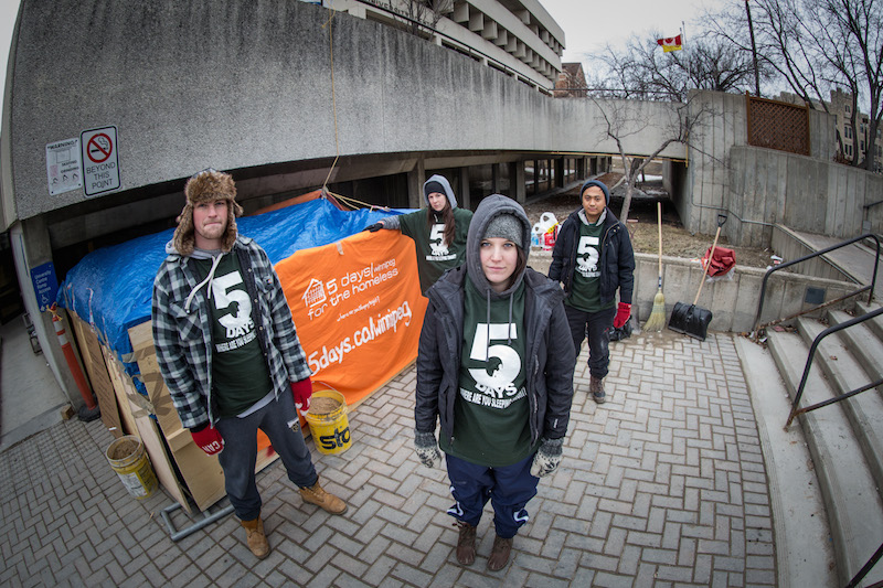 Four students stand outside their makeshift home made of cardboard and tarps