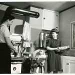 A photograph of a family inside the kitchen of a home in Veterans' Village at the University of Manitoba Campus. A man is standing at the stove with a pot, an infant is in a high chair, and a woman appears to be holding a loaf of bread and a bag in her hands. Photograph is dated as having been taken around 1948 by the National Film Board of Canada. Source: University Relations & Information Office fonds