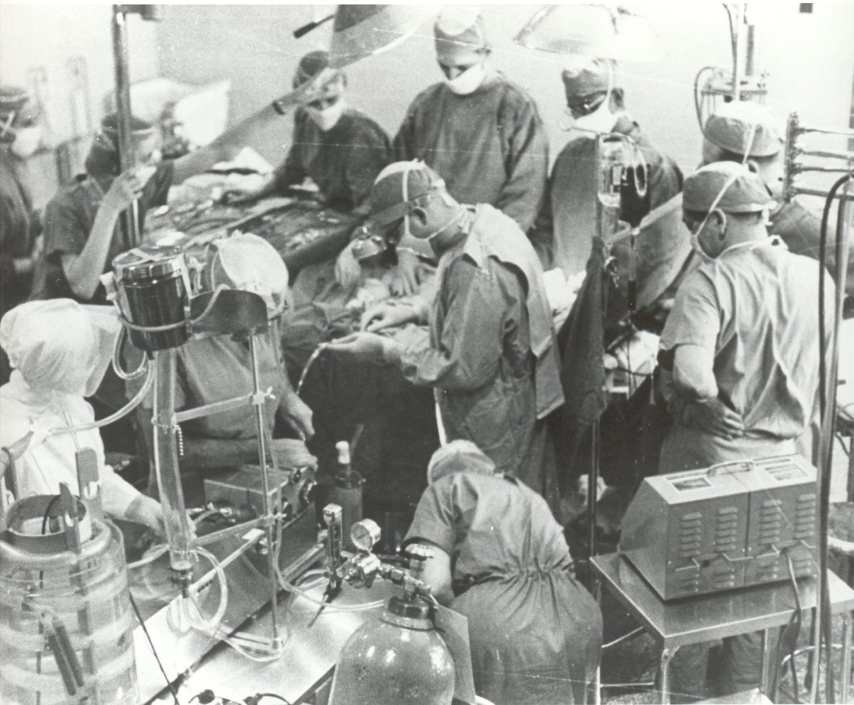 Black and white photo of 11 people in an operating room with medical equipment. The people are watching the surgery or doing tasks.