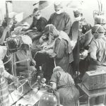 Black and white photo of 11 people in an operating room with medical equipment. The people are watching the surgery or doing tasks.