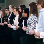 Male and female students wearing black and white hold white candles which are lit.