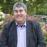 Todd Duhamel sitting in front of flowers and shrubs on the campus of the University of Manitoba