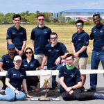 Nine students wearing team branded navy blue polo shirts stand/sit posing facing the camera. They are surrounding their aircraft build which is white and about 8 feet in wing span. In front of the plane are three award plaques.