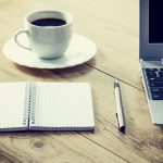 Cup of coffee on a saucer sits on a wooden tabletop beside a laptop, pen, and open notebook.