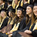 A group of UM grads sit in a row at Convocation. They wear black caps and gowns with golden hood linings. The graduate in the middle smiles at the camera as all hold up their degree parchments.