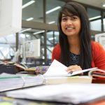 Woman with dark hair smiling in the Asper School of Business Drake Building. She has books and papers infront of her that are out of focus.