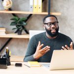 A man wearing glasses and a green sweater is smiling at the computer and talking with his hands.
