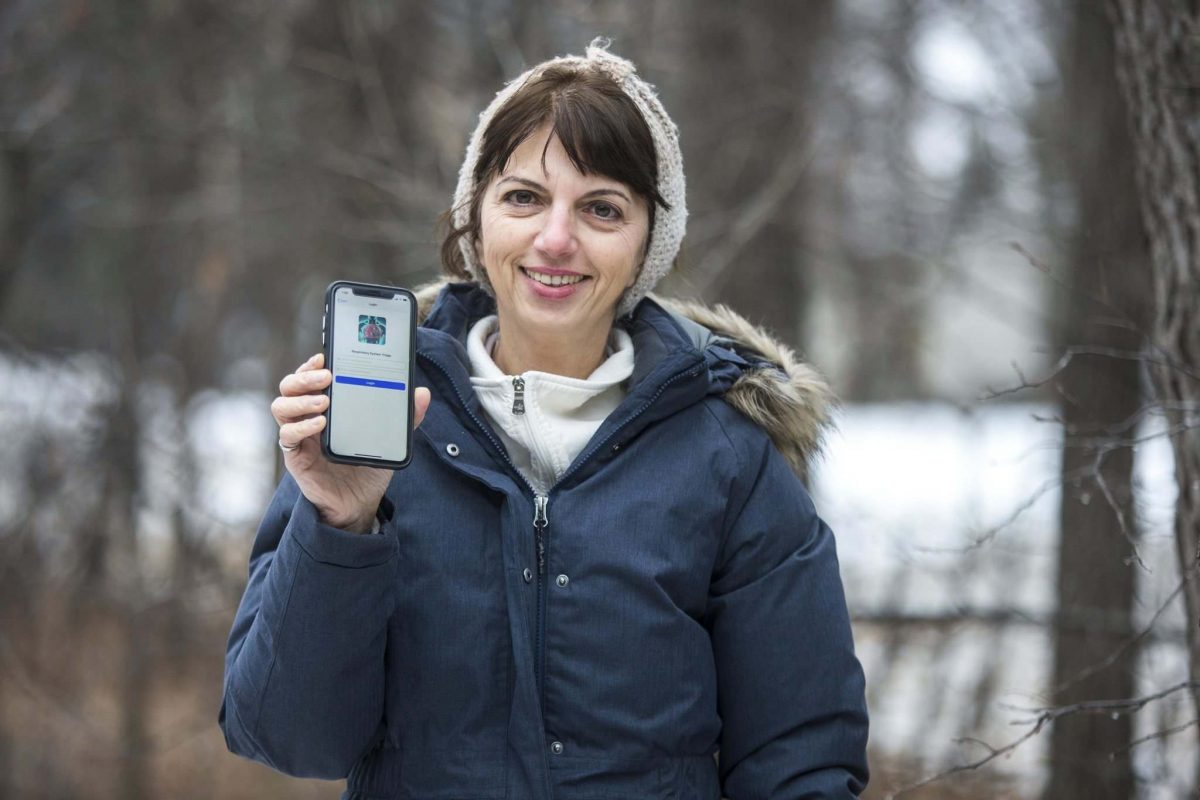 Doctor Zahra Moussavi standing outside smiling and posing for the camera while holding up a smartphone.