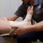 The arms and upper body of a physiotherapy student are seen. The student is taping a woman's bare foot and ankle with athletic tape.