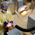 A dental student wearing personal protective equipment performs a dental procedure on a patient while a plastic funnel attached to plastic tubing vacuums aerosols away from the patient's chin area.
