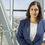 Woman in professional clothing stands in front of bank of windows with the Winnipeg cityscape in view behind her.