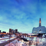 Canadian Museum of Human Rights building with a blue sky background, with a snowy foreground and the downtown skyline in the distance.