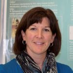 Woman with shoulder length dark brown hair with bangs. Wearing a blue sweater with a dark coloured scarf around her neck.