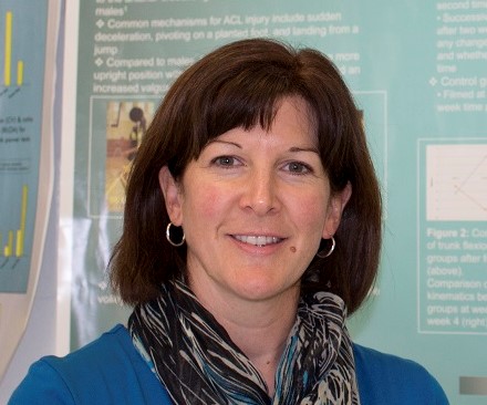 Woman with shoulder length dark brown hair with bangs. Wearing a blue sweater with a dark coloured scarf around her neck.