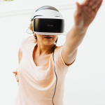 A standing woman wearing a virtual-reality headset stretches one arm upward in front of her.
