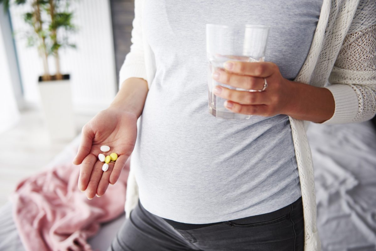 Pregnant woman wearing a light-colored top, holding out her hand to display a small pile of pills, with her other hand resting gently on her belly. The background is softly blurred, drawing attention to the pills and the woman's gesture.