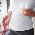 Pregnant woman wearing a light-colored top, holding out her hand to display a small pile of pills, with her other hand resting gently on her belly. The background is softly blurred, drawing attention to the pills and the woman's gesture.