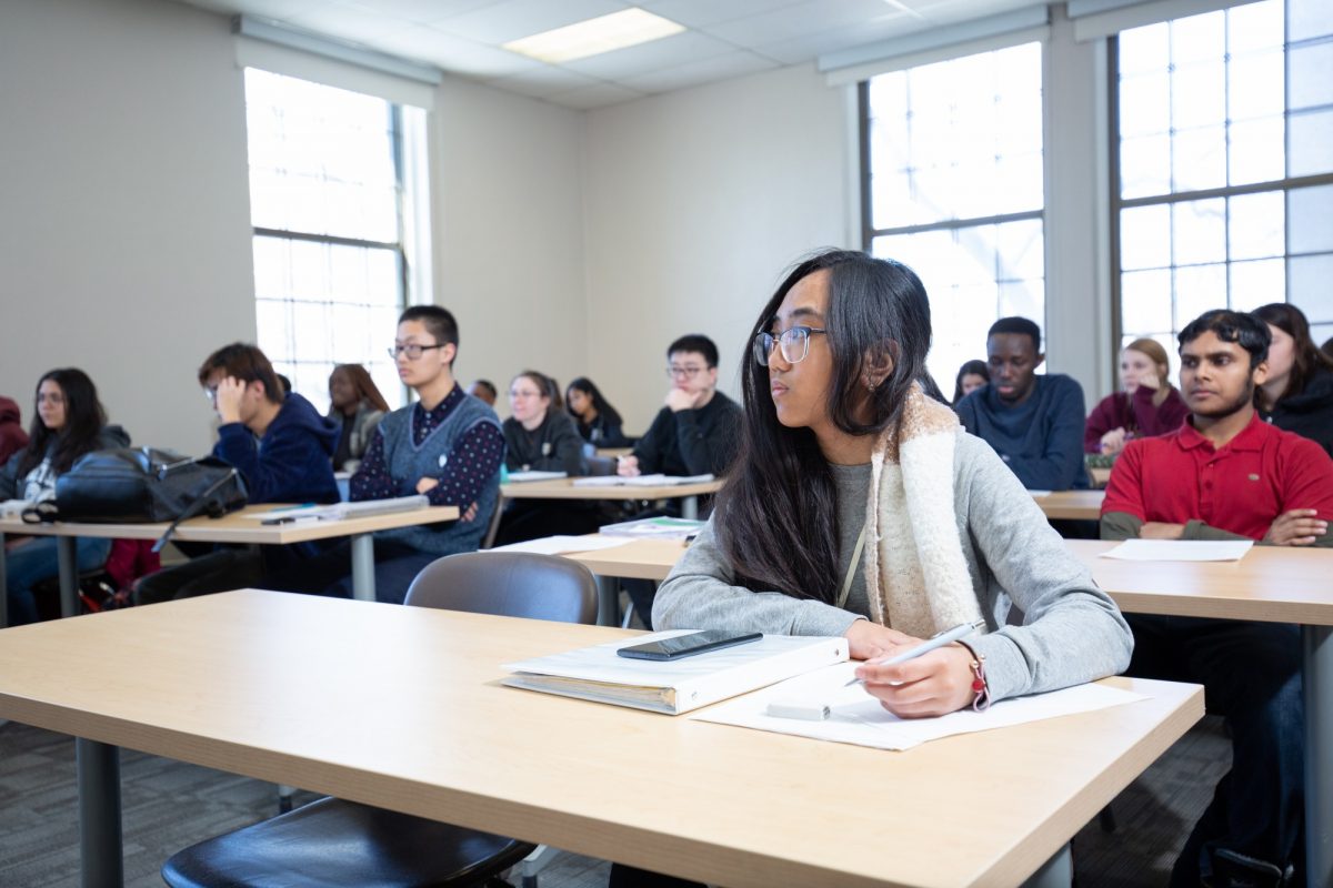 A group of students listen to the instructor in class. A young female student with long brown hair and glasses takes notes.