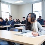 A group of students listen to the instructor in class. A young female student with long brown hair and glasses takes notes.