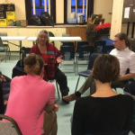 A group of people sitting on chairs in a circle, listening to Audrey Logan speak.