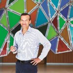 Dr. Prabhat Jha stands in front of a colourful wall decoration.
