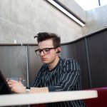 A white guy wearing glasses and a black shirt with white stripes works at a computer on campus.