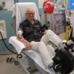 A man sitting upright in a hospital chair with his legs stretched in front of him pedals a stationary cycling wheel while receiving dialysis.
