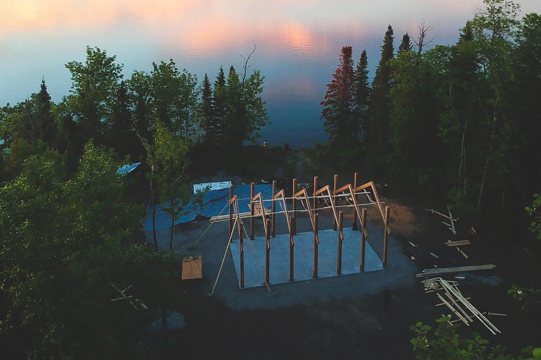 An aerial photo of a building being constructed on treed land near a lake.