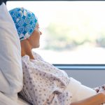 A female cancer patient looks out a window while propped up in a hospital bed.