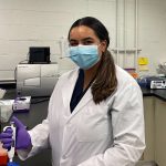 Navi Mann in a lab. She is wearing a lab coat and face mask.