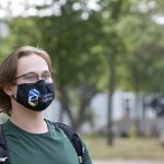 A student wearing a green shirt and a black mask with the University of Manitoba logo on it stands among the trees on the Fort Garry Campus.
