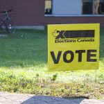 A yellow Elections Canada sign with the word VOTE on it in bold letters placed in the grass along a sidewalk.