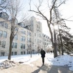 A student walks alone on campus during the winter, heading towards a white brick building.