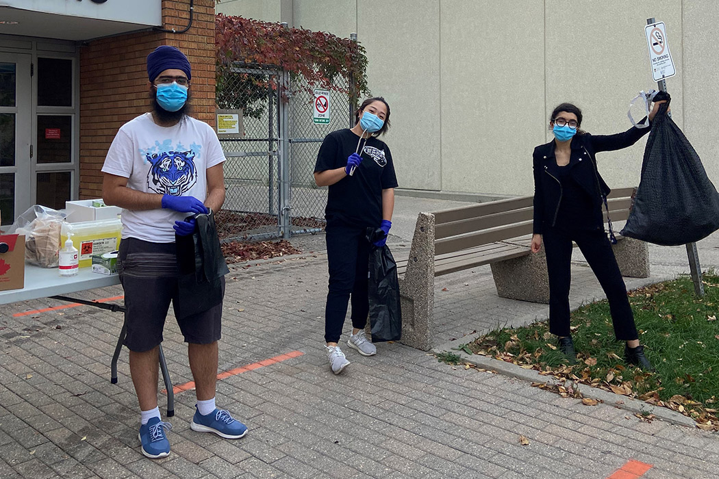 Three students stand outside. One student holds up a filled garbage bag.