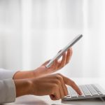 Hands typing on keyboard and looking at mobile phone.
