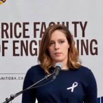 Avery Edwards stands with a netural expression while standing at a podium with a microphone. She is wearing a white ribbon on her right lapel.