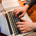 a student's hands type on a laptop. School books sit on the desk in the background.