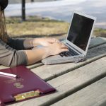 A red UM clipboard sits on a picnic table in front of a person typing on a laptop computer.