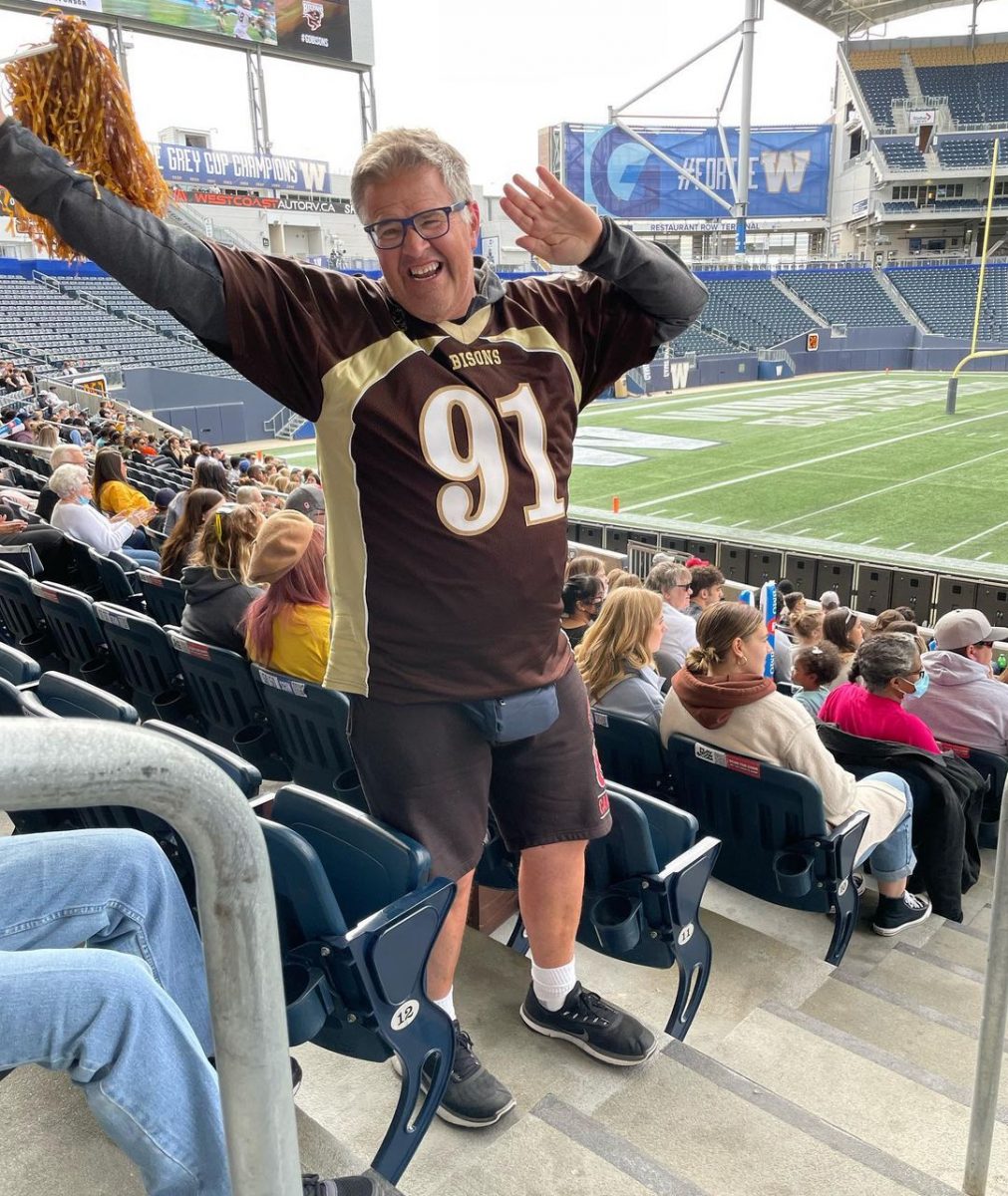 Man in brown and gold sports jersey strikes a dancing pose in the stands of a football stadium