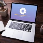 A laptop sits on a desk with a program update loading on its screen. It is surrounded by an open notebook, a cup of coffee and a small potted plant