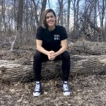 Asper alum Leanne Kisil wearing all black sitting on a log in the forest smiling.