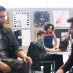 Two people seated in the foreground, two people looking at a computer screen in the background.