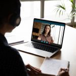 person with headphones on looking at laptop screen where woman is online chatting