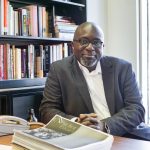 Professor Barrington Walker leaning on his desk with bookshelf behind.