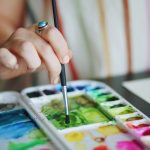 A hand holding a paintbrush grabs green paint off a colourful palette