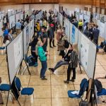 side angle shot of students standing in front of their research posters in an auditorium