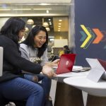 Two students look at a red laptop on a table in Campo. They are excited about what they see on the screen.