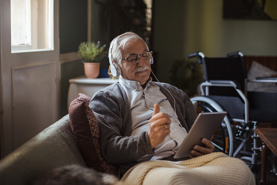 An elderly man looks at a tablet while wearing headphones. A wheelchair is in the background